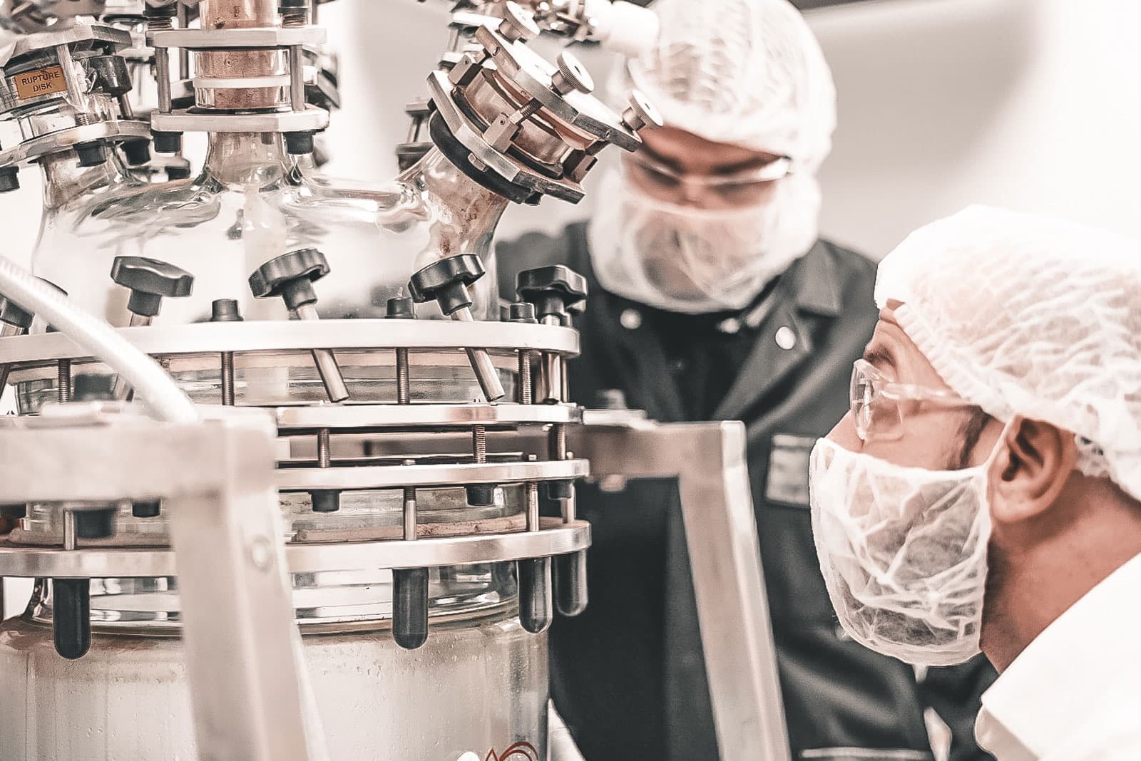Due operatori in camice e cuffia sterile al lavoro su un reattore in vetro del laboratorio